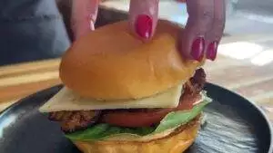 Hand placing bun on a delicious grilled chicken sandwich with cheese, lettuce, and tomato on a black plate.