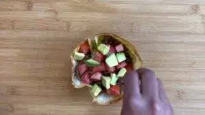 Taco salad with avocado and tomato pieces on a wooden board, hand arranging toppings for a fresh meal.