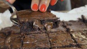 Close-up of a gooey chocolate brownie slice with a glossy top, held by a hand with red nails, tasty dessert.