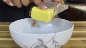 Person placing butter into a white bowl on a wooden countertop.