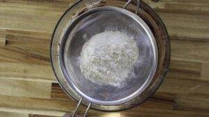 Sifting flour into a mixing bowl on a wooden surface for baking preparation.