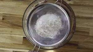 Sifting flour into a mixing bowl on a wooden surface for baking preparation.
