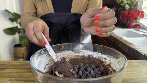 Person mixing brownie batter with chocolate chips in a glass bowl, wearing orange nail polish.