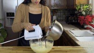 Woman using a hand mixer while pouring ingredient into bowl in kitchen.