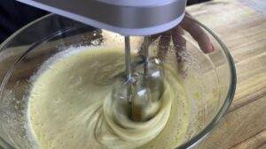 Mixing creamy cake batter with an electric mixer in a glass bowl on a wooden table. Baking process close-up.