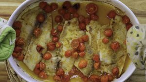 Baked fish with cherry tomatoes in a white dish, displayed on wooden table, garnished with spices.