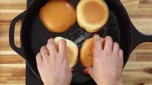 Toasting burger buns in a skillet with hands turning them gently.