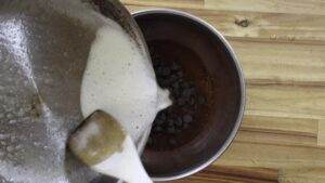 Pouring cream into a bowl of chocolate chips, wooden spoon on the side, on a wooden table.