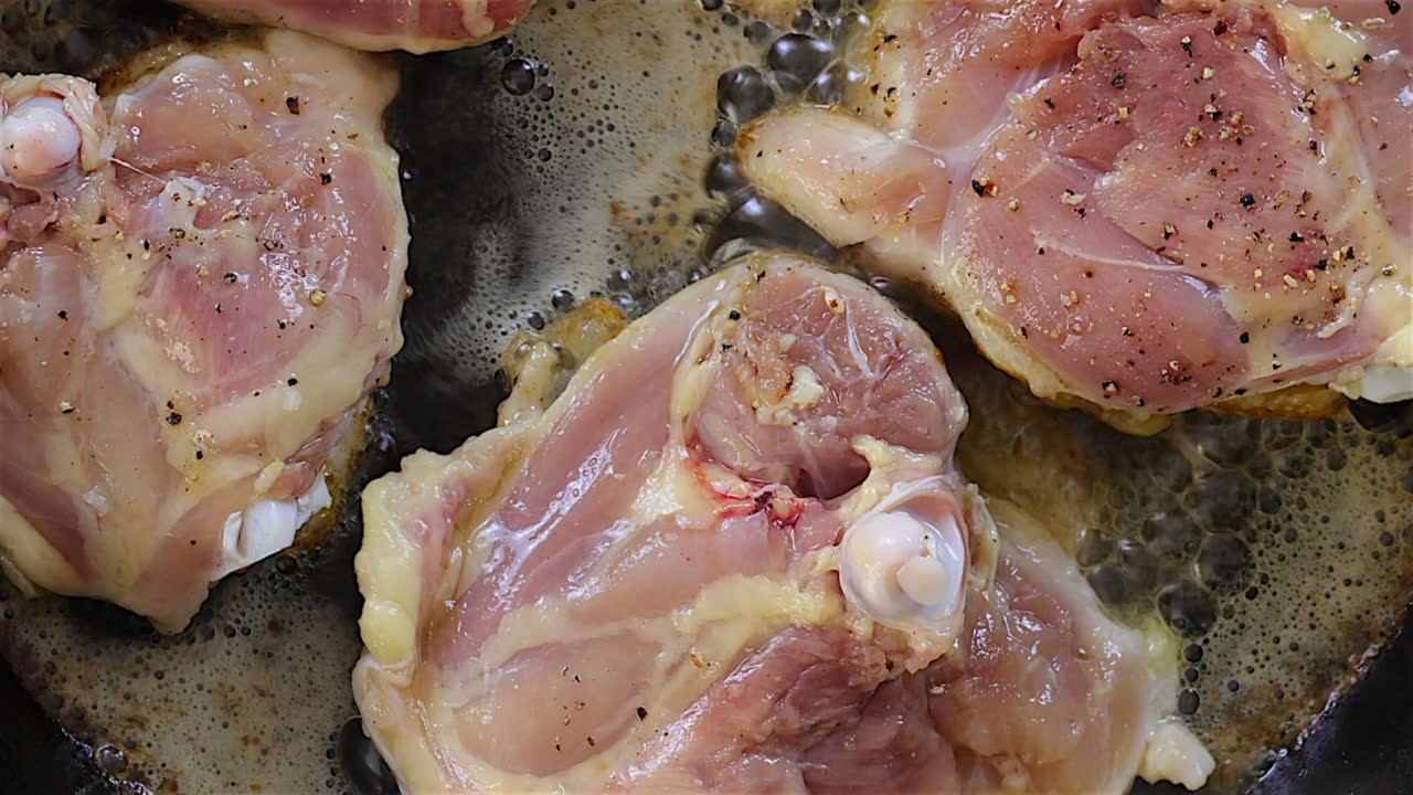 Raw chicken thighs cooking in a skillet with sizzling oil and seasoning.
