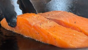 Close-up of seasoned salmon fillets being cooked in a pan with tongs, highlighting texture and spices.