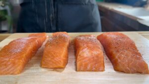 Fresh salmon fillets being seasoned with salt and pepper on a wooden board, ready for cooking.