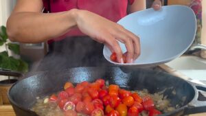 Person adding cherry tomatoes to a sizzling cast iron skillet with sautéed onions. Home cooking in action.