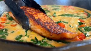 Seared salmon being added to a creamy sauce with spinach and cherry tomatoes in a skillet.