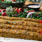 Grilled kebabs with herbs on flatbread, served with fresh tomato salad and a side of sauce.