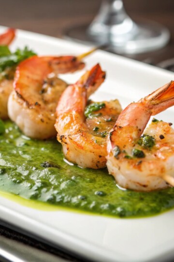 Grilled shrimp skewers on basil sauce served on a white plate, garnished with fresh herbs.