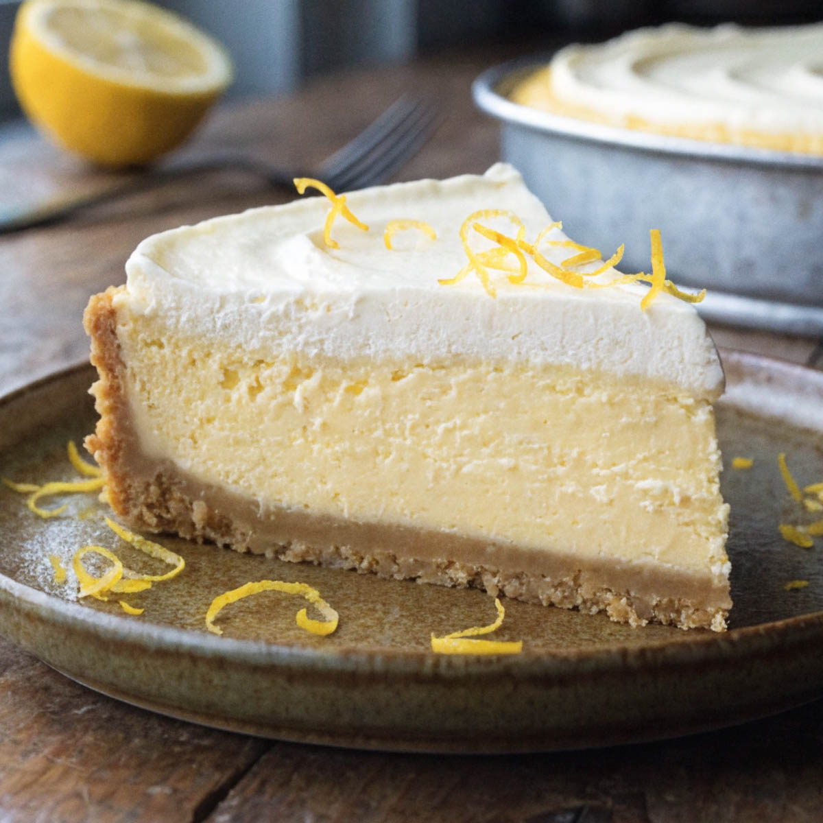 Slice of creamy no bake lemon mascarpone cheesecake with zest garnish on a ceramic plate, half lemon and fork in background.