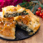 Freshly baked spinach and feta pie with golden phyllo crust, served on a plate, cherry tomatoes in background.