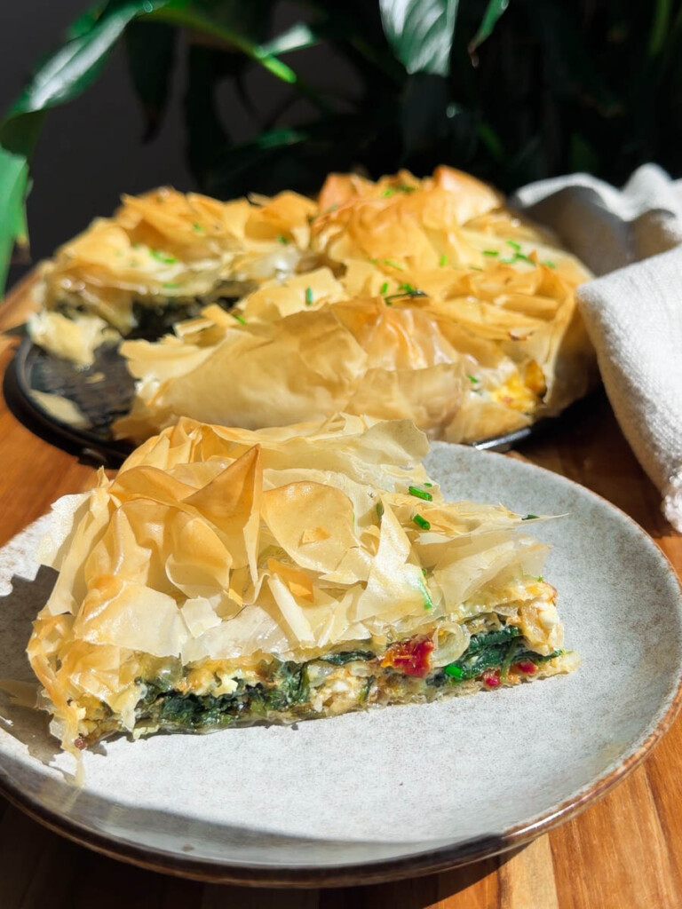 Slice of Mediterranean phyllo pie with crispy filo pastry on a plate, garnished with herbs.
