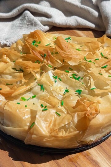 Mediterranean phyllo pie with crispy phyllo layers, topped with green chives, on a wooden board and cloth backdrop.