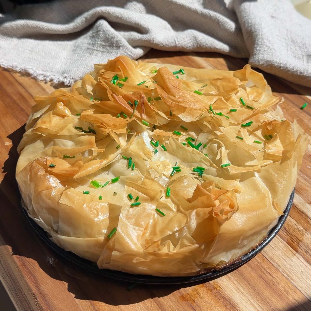 Mediterranean phyllo pie with crispy phyllo layers, topped with green chives, on a wooden board and cloth backdrop.