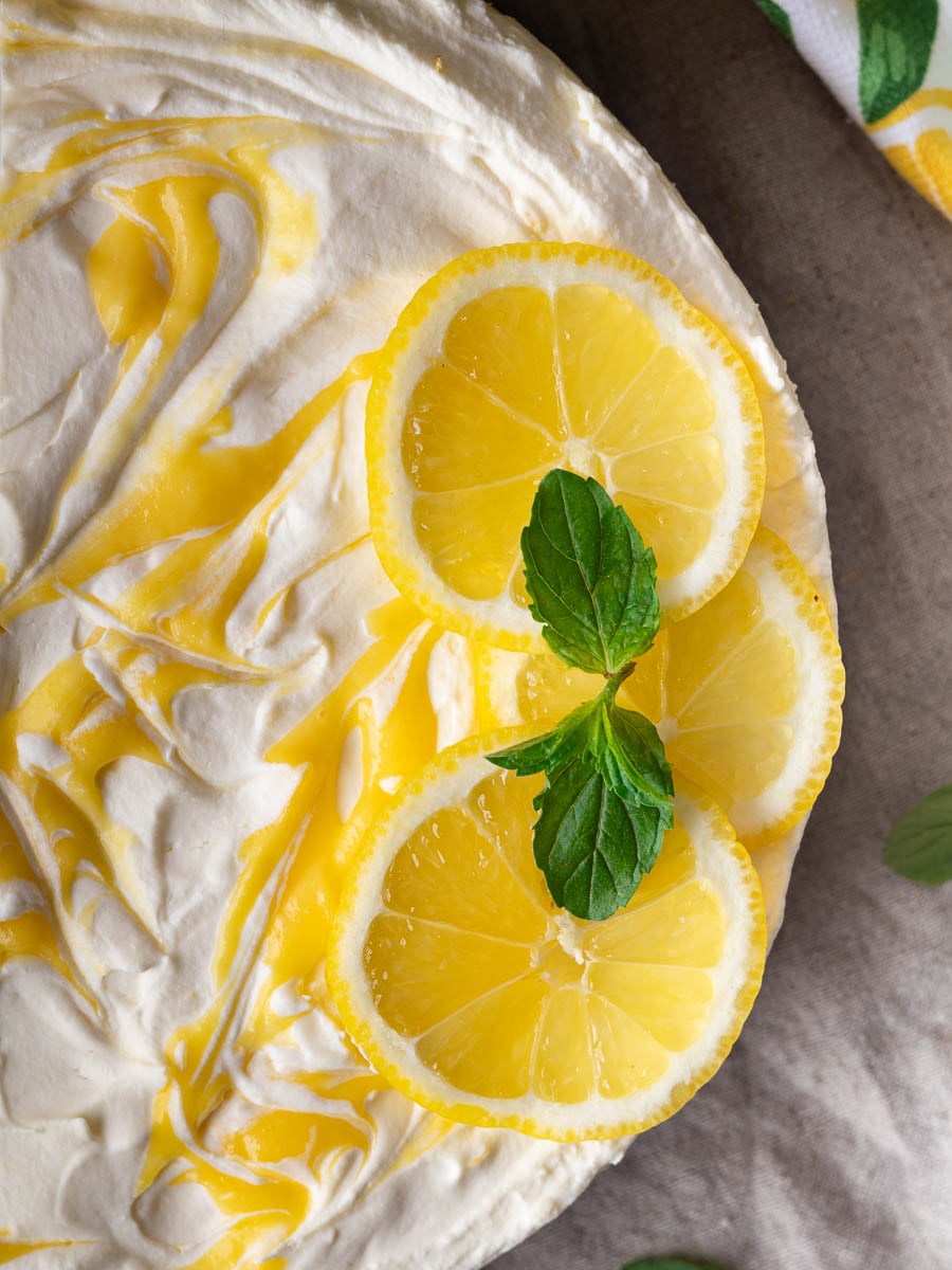 No-bake lemon mascarpone cheesecake with whipped cream, lemon slices, and mint leaves on top, on a brown cloth background.