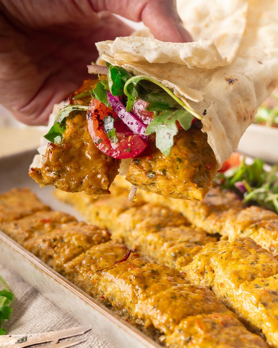 Hand holding sheet pan chicken kebabs in the oven with fresh vegetables over a tray of kebabs.