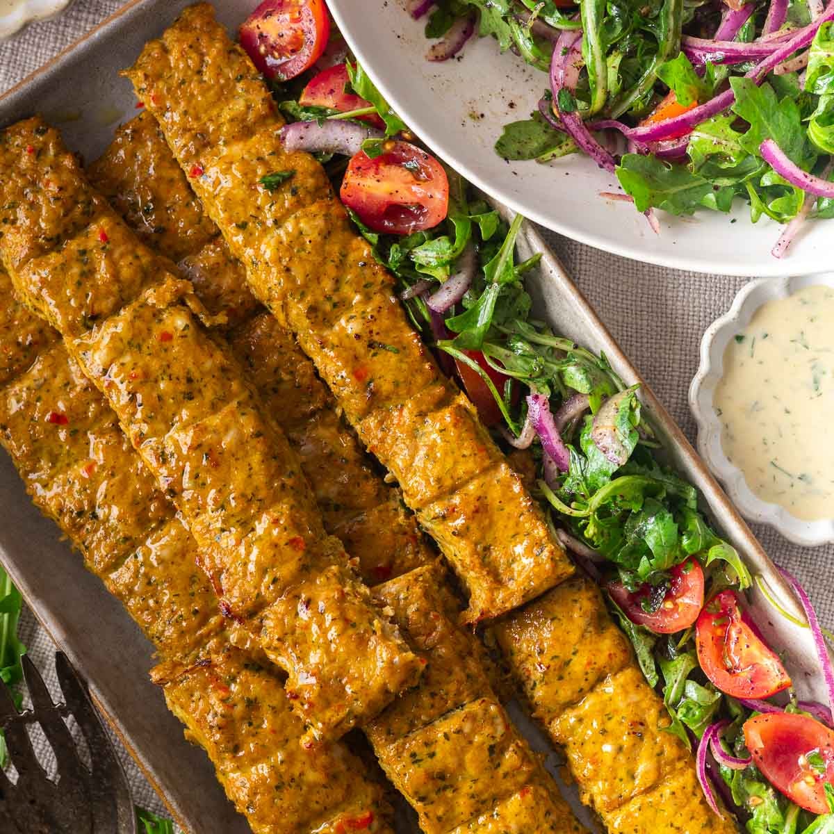 Sheet pan chicken kebabs in the oven with salad and dipping sauce on a platter, featuring tomatoes and arugula.