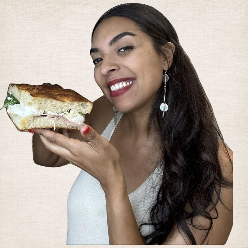 Woman smiling, holding a large sandwich with fillings, wearing earrings and a sleeveless top.