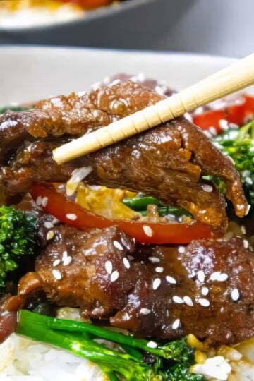 Beef stir-fry with broccoli, red peppers, and sesame seeds over rice, held with chopsticks. Asian cuisine in a bowl.