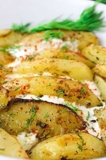 Roasted potato wedges with herbs and cream, garnished with fresh dill in a white bowl.