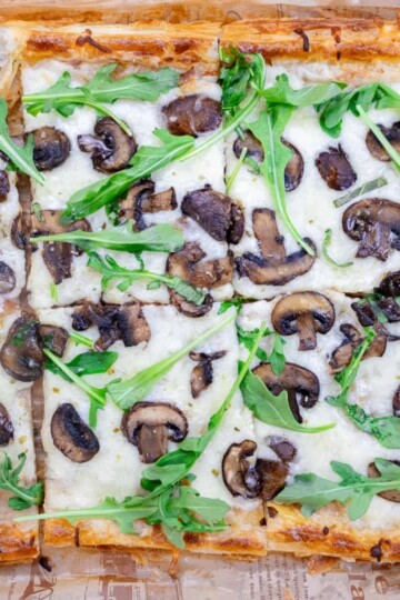 Flatbread pizza topped with mushrooms, arugula, and melted cheese, cut into squares on a wooden board.