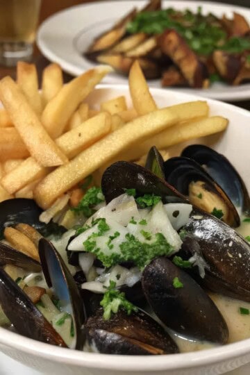 Bowl of steamed mussels in creamy sauce with golden fries, garnished with parsley; comfort seafood dish.