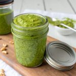 Green pistachio pesto sauce in a glass jar on a wooden board with peanuts and a spoon nearby.