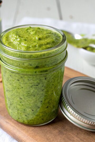 Green pistachio pesto sauce in a glass jar on a wooden board with peanuts and a spoon nearby.