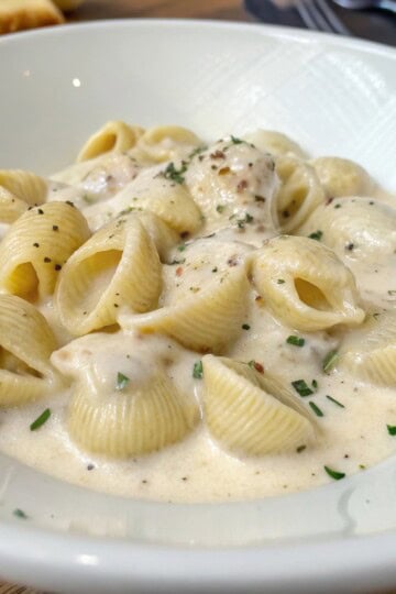 Creamy pasta shells in rich white sauce, garnished with herbs and pepper in a white bowl.