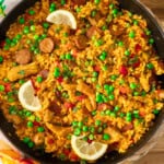 Pan of vibrant paella with saffron rice, peas, chorizo, chicken, red peppers, and lemon wedges.