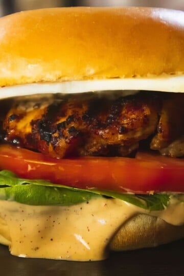 Close-up of a grilled chicken sandwich with lettuce, tomato, cheese, and sauce on a toasted bun.