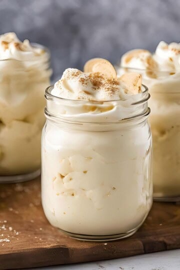 Creamy eggnog banana pudding jars topped with whipped cream and banana slices on a wooden board.