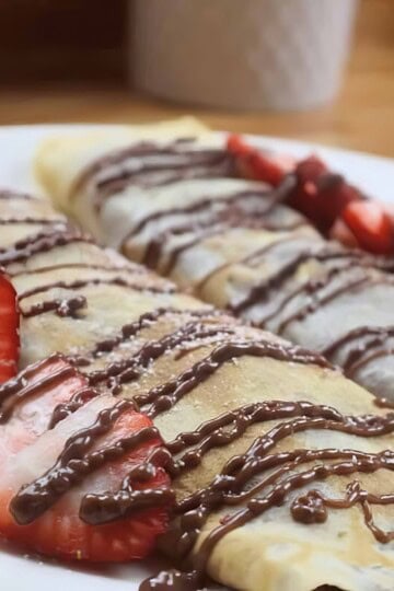 Crepes topped with fresh strawberries and a drizzle of chocolate sauce on a white plate.