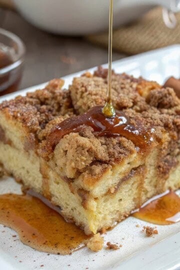 Close-up of cinnamon coffee cake drizzled with honey on a white plate, garnished with cinnamon sticks.