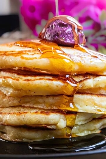 Stack of fluffy pancakes drizzled with syrup, topped with a purple ice cream scoop, on a black plate.