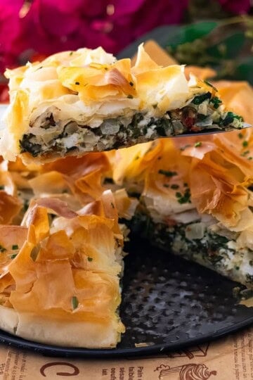 Freshly baked spinach pie with crispy phyllo layers, served on a platter with a slice lifted and vibrant floral backdrop.