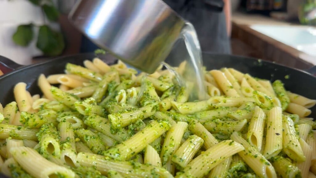 Pouring pasta water into pesto pasta in a skillet, creating a delicious pesto pasta.