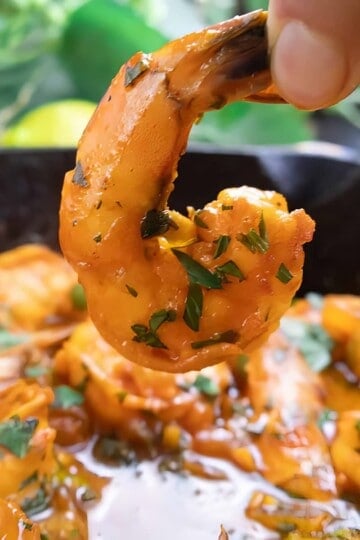 Close-up of a cooked, seasoned shrimp held above a pan, garnished with fresh herbs.