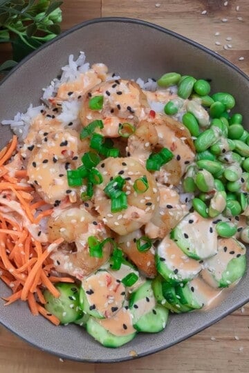 Shrimp poke bowl with rice, edamame, carrots, and cucumbers, garnished with sesame seeds and green onions.