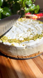 Knife slicing pistachio-topped cheesecake on wooden board with leafy background.