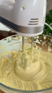 Electric hand mixer blending creamy batter in a glass bowl.