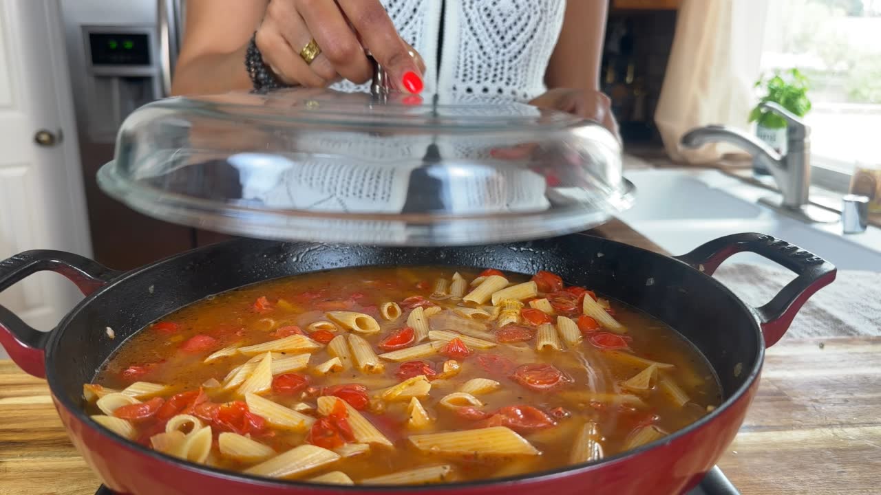 Cooking penne pasta with tomatoes in a skillet, person lifting lid in a bright kitchen setting.