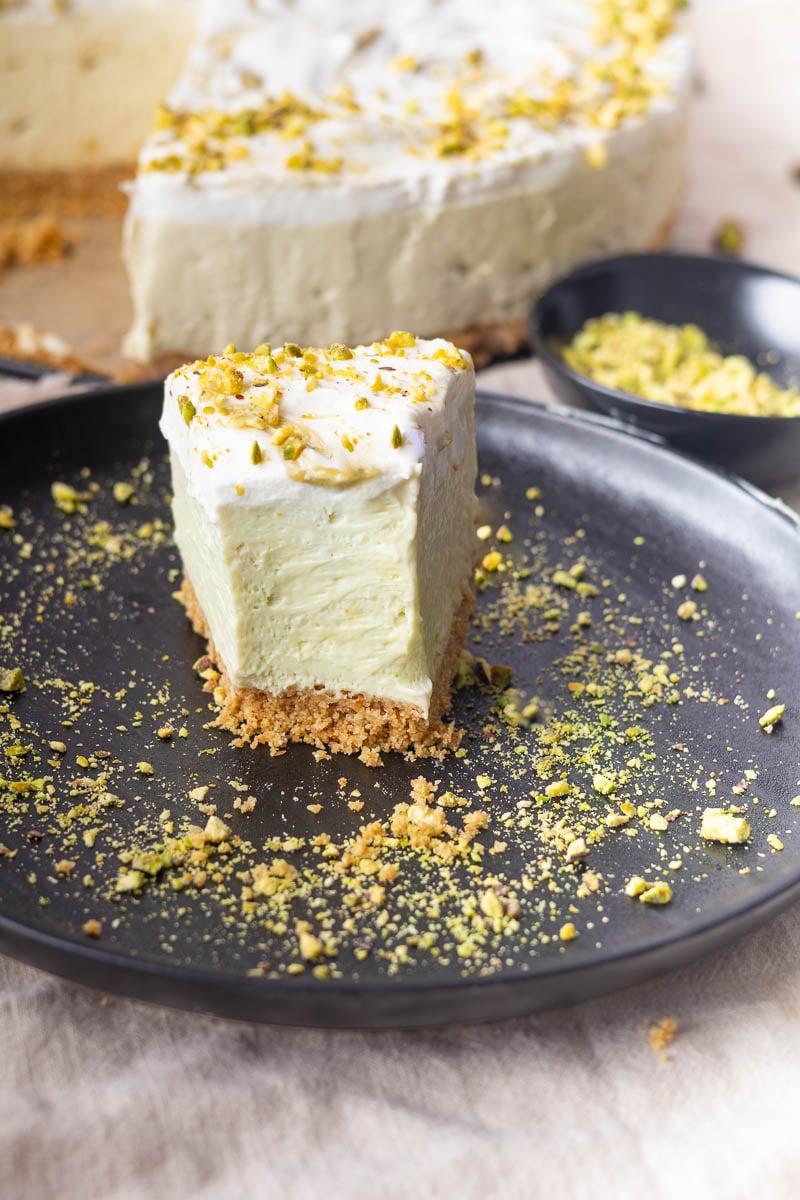 Slice of no-bake pistachio mascarpone cheesecake with whipped cream on plate, garnished with crushed pistachios.