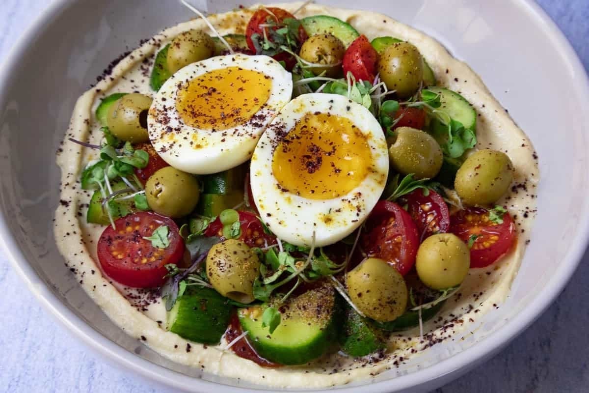Mediterranean Hummus Bowl topped with soft-boiled eggs, cherry tomatoes, olives, and greens.